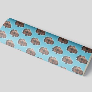 May include: A roll of wrapping paper with a repeating pattern of brown faces on a light blue background. The faces have dark hair and are in a cartoon style. The roll is slightly curved.