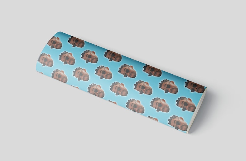 May include: A roll of wrapping paper with a repeating pattern of brown faces on a light blue background. The faces have dark hair and are in a cartoon style. The roll is slightly curved.