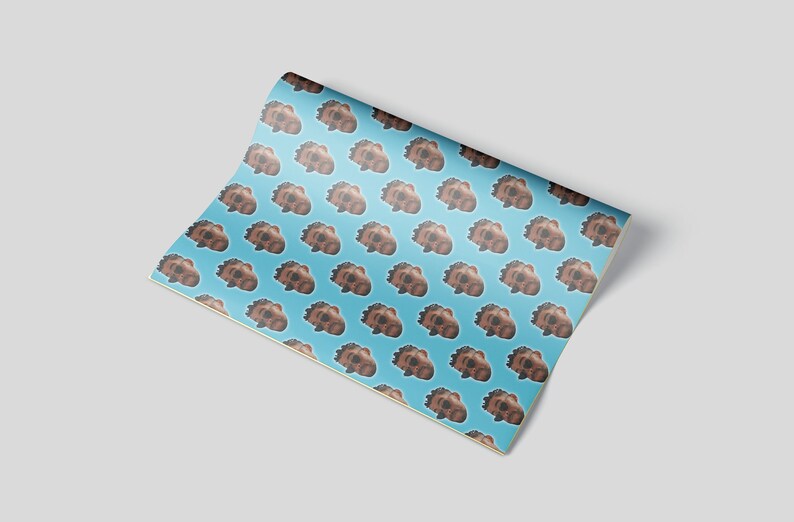 May include: A roll of wrapping paper with a repeating pattern of brown cartoon faces on a light blue background. The paper is partially unrolled, revealing the pattern.
