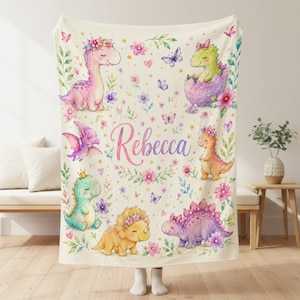 May include: A cream-colored throw blanket featuring a watercolor print of various colorful dinosaurs, including a pink long-neck, a green hatchling, and a purple stegosaurus, decorated with flowers, butterflies, and the name Rebecca in pink script.