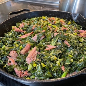 May include: A cast iron skillet filled with collard greens and pieces of ham. The greens are dark green and the ham is pink.