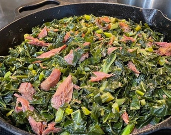 May include: A cast iron skillet filled with collard greens and pieces of ham. The greens are dark green and the ham is pink.