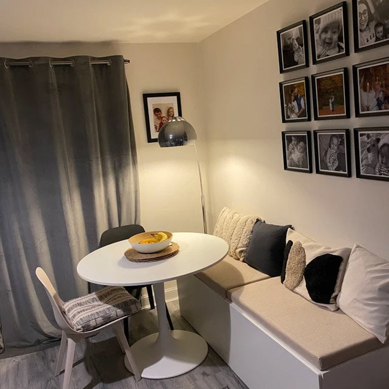 May include: A white oval dining table with a central pedestal base, accompanied by a single chair with a patterned cushion, a built-in bench with beige cushions and decorative pillows, a chrome floor lamp, and framed wall photographs.