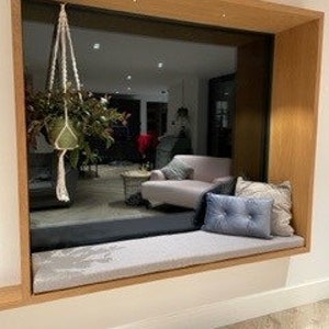 May include: A window seat with a gray cushion and two blue throw pillows. The window is framed in wood and has a view of a room with a couch and a coffee table.