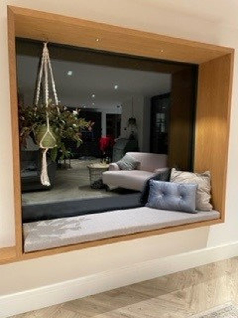 May include: A window seat with a gray cushion and two blue throw pillows. The window is framed in wood and has a view of a room with a couch and a coffee table.