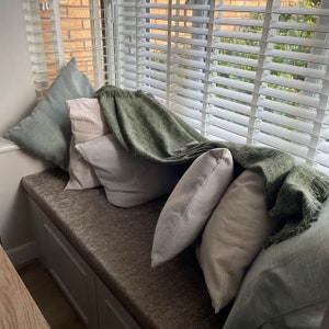 May include: A window seat cushion upholstered in a textured brown fabric, featuring a collection of square throw pillows in shades of sage green and off-white, draped with a fringed olive green woven throw blanket against white window blinds.