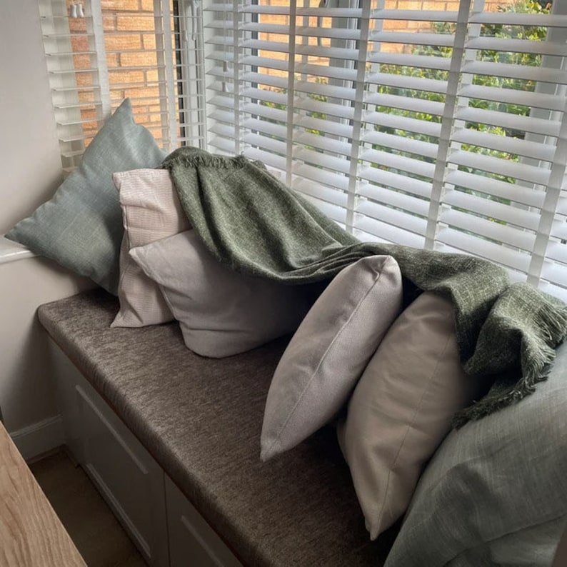 May include: A window seat cushion upholstered in a textured brown fabric, featuring a collection of square throw pillows in shades of sage green and off-white, draped with a fringed olive green woven throw blanket against white window blinds.