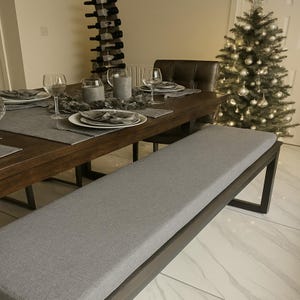 May include: A long dining bench with a dark wood frame and a light gray upholstered seat cushion. The bench is positioned in front of a dark wood dining table set with plates, glasses, and gray napkins, with a Christmas tree nearby.