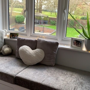 May include: A window seat featuring a gray textured cushion, a square gray pillow with pom-poms, a heart-shaped cream pillow, and a rectangular gray pillow. An aloe vera plant in a gray pot and two small framed photographs sit on the window sill.