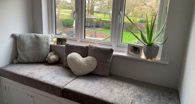May include: A window seat featuring a gray textured cushion, a square gray pillow with pom-poms, a heart-shaped cream pillow, and a rectangular gray pillow. An aloe vera plant in a gray pot and two small framed photographs sit on the window sill.