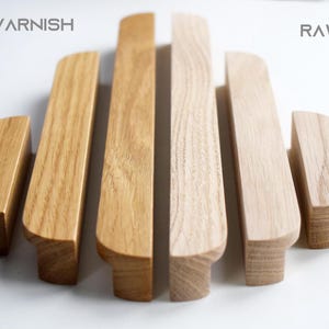 May include: A set of eight wooden drawer pulls in various sizes, shown in both raw wood and clear varnish finishes. The pulls are all rounded on the edges and have a simple, modern design. The text "CLEAR VARNISH" and "RAW WOOD" is displayed above the pulls.