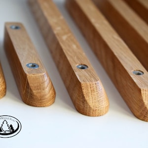 Pu&ograve; includere: Cinque maniglie per cassetti in legno con bordi arrotondati e finitura in legno naturale. Ogni maniglia &egrave; dotata di due viti argentate per l'installazione. Le maniglie sono disposte in fila su una superficie bianca.