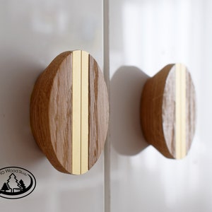 May include: Two wooden cabinet knobs with a light brown finish and a vertical gold-coloured stripe down the centre. The knobs are mounted on a white surface, casting a shadow.