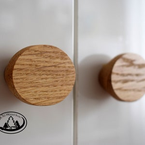 May include: Two wooden knobs with a natural wood grain finish. The knobs are round and have a slightly raised edge. The knobs are mounted on a white surface.