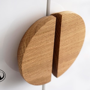 Op de afbeelding: Twee houten kastdeurgrepen, elk in de vorm van een halve cirkel, zijn bevestigd aan een wit oppervlak. De grepen zijn gemaakt van lichtbruin hout met een natuurlijk houtnerfpatroon. De grepen zijn met een metalen staaf aan het oppervlak bevestigd.