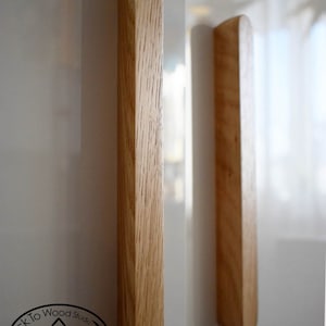 Pu&ograve; includere: Due maniglie per armadi in legno con forma rettangolare arrotondata. Le maniglie sono in legno marrone chiaro con una venatura naturale. Le maniglie sono montate su una superficie bianca.