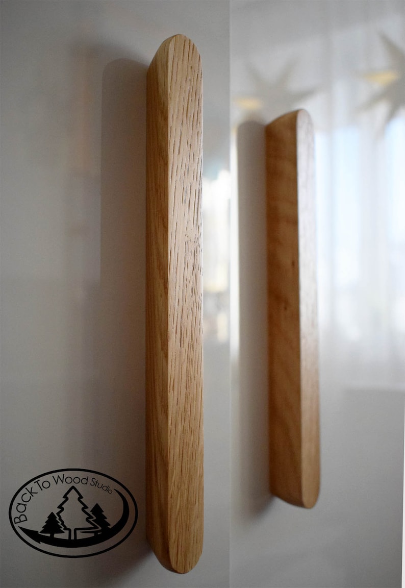 Pu&ograve; includere: Due maniglie per armadi in legno con forma rettangolare arrotondata. Le maniglie sono in legno marrone chiaro con una venatura naturale. Le maniglie sono montate su una superficie bianca.