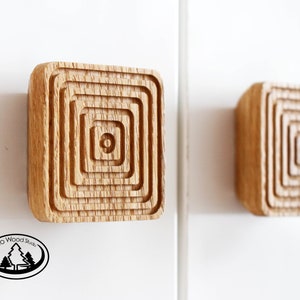 Könnte beinhalten: Zwei quadratische Holzknauf für Schränke mit konzentrischem Kreisdesign. Die Knäufe sind aus hellem braunem Holz gefertigt und haben eine glatte Oberfläche.