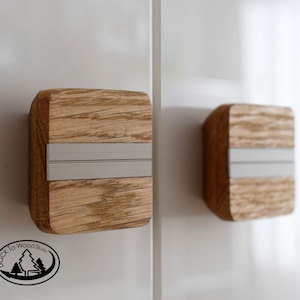 Peut inclure: Deux boutons de cabinet carrés en bois avec un incrustation en métal argenté. Les boutons ont un motif de grain de bois et sont montés sur une surface blanche.