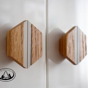Puede incluir: Dos pomos hexagonales de madera para armario con una franja blanca en el centro. Los pomos están montados en una superficie blanca.