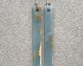May include: A pair of long, rectangular drop earrings featuring a smooth, matte blue-grey stone surface with natural, irregular golden-brown veining. Each earring is suspended from a thin, silver-toned metal wire hook through a small circular hole.