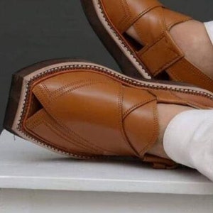 May include: Close-up of brown leather sandals with a strap and open design. The sandals have a stitched sole and a dark brown sole. The leather appears smooth and the sandals are resting on a white surface.