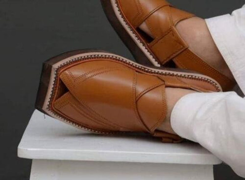 May include: Close-up of brown leather sandals with a strap and open design. The sandals have a stitched sole and a dark brown sole. The leather appears smooth and the sandals are resting on a white surface.