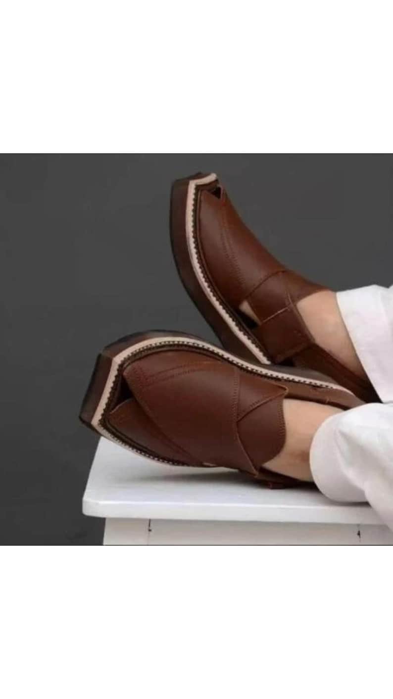 May include: A pair of brown leather Peshawari sandals. The sandals have a rounded toe and a strap across the top. The soles are a lighter color with a white stitched border. The sandals are on a white surface.