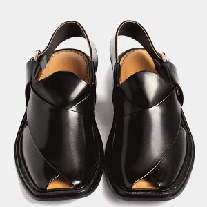May include: A pair of black leather Peshawari sandals. These open-toe sandals feature a distinctive crisscross design across the top, a slingback strap, and a square toe. The interior is a light tan color, and the soles are black.