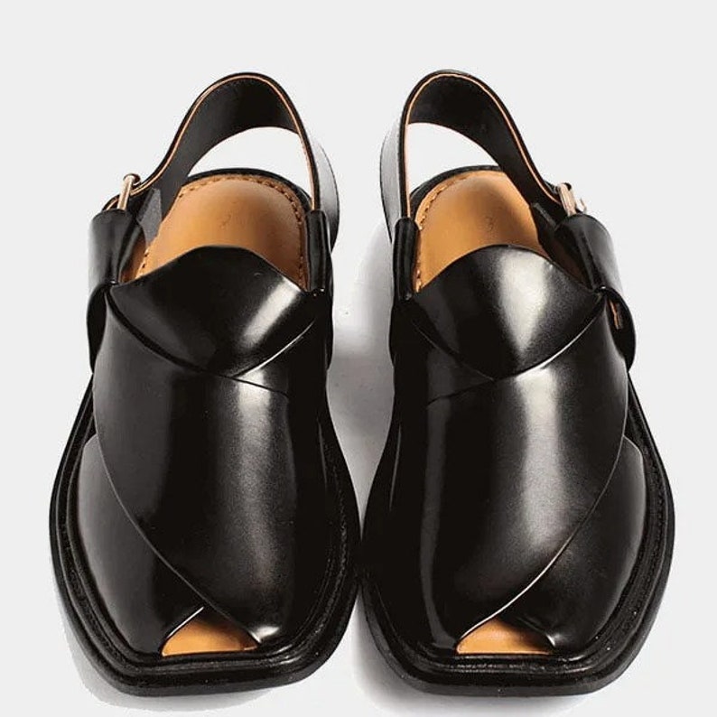May include: A pair of black leather Peshawari sandals. These open-toe sandals feature a distinctive crisscross design across the top, a slingback strap, and a square toe. The interior is a light tan color, and the soles are black.