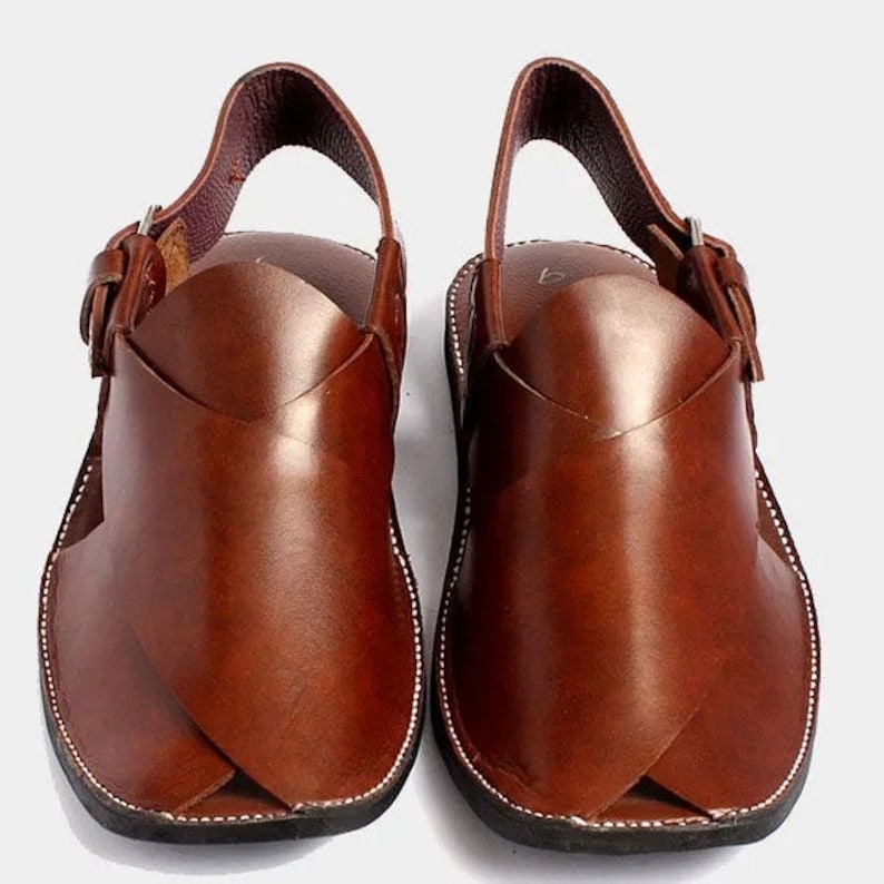Puede incluir: Un par de sandalias de cuero marr&oacute;n con correa trasera y cierre de hebilla. Las sandalias tienen un dise&ntilde;o &uacute;nico con paneles de cuero superpuestos en la parte delantera. Costuras blancas delinean la suela.