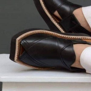 May include: Black leather sandals with a thick sole. The sandals have a stitched design and a light brown trim around the sole. The image shows the sandals on a white surface.