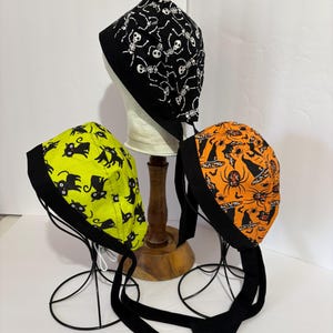 May include: Three scrub caps featuring Halloween patterns. One cap is black with white skeletons, one is lime green with black cats, and one is orange with black spiders and witch hats. Each cap has a solid black fabric band and long tie straps.