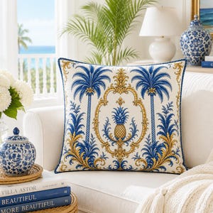Könnte beinhalten: Ein quadratisches Zierkissen mit cremefarbenem Hintergrund, verziert mit einem symmetrischen Muster aus blauen Palmen und einem zentralen goldenen Ananas-Motiv. Das Design umfasst filigrane goldene Schnörkel und eine dunkelblaue Paspelierung.
