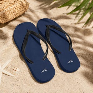 May include: A pair of navy blue flip-flops with black thong straps. Each sandal features a smooth, solid-colored footbed and a small white graphic of a stylized wave near the heel. The design is simple, featuring a flat sole and a classic thong construction.