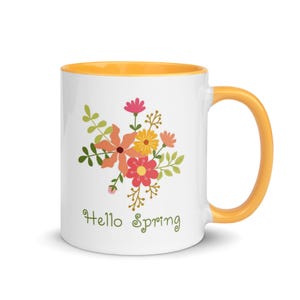 May include: White ceramic mug with a yellow interior and handle. The mug features a floral design in shades of orange, pink, and yellow, with green leaves. The words "Hello Spring" are written in green script below the flowers.