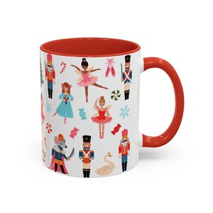 May include: White ceramic mug with a red interior and handle. The mug is decorated with colorful illustrations of nutcrackers, ballerinas, candy canes, and snowflakes, creating a festive holiday theme.