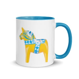 May include: White ceramic mug with a blue handle and interior. The mug features a yellow Dala horse design with blue and white accents. The mug is a standard size for coffee or tea.