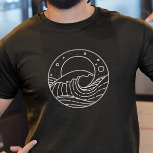 K&ouml;nnte beinhalten: Ein Mann tr&auml;gt ein schwarzes T-Shirt mit einem wei&szlig;en Grafikdruck einer Welle und Sternen in einem Kreis. Er tr&auml;gt eine blaue Baseballkappe und eine blaue Jeans.