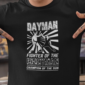 May include: Black t-shirt with a white graphic design featuring a stylized image of two figures fighting. The text "DAYMAN", "FIGHTER OF THE NIGHTMAN", and "CHAMPION OF THE SUN" is printed on the shirt.
