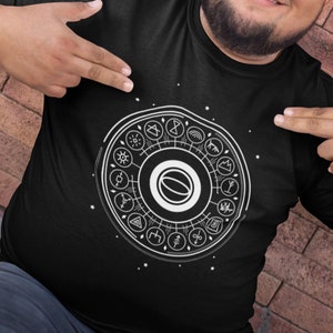 May include: A black t-shirt with a white graphic design of a circle with various symbols inside. The circle is surrounded by stars. The person wearing the shirt is smiling and pointing at the graphic.