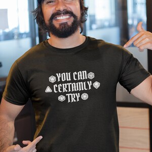 May include: A black t-shirt with the text "YOU CAN CERTAINLY TRY" and a graphic of dice. The person wearing the shirt is smiling and pointing at the shirt.