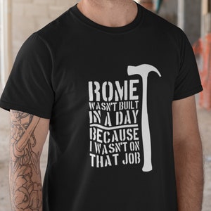 May include: A black t-shirt with a white graphic design that says "ROME WASN'T BUILT IN A DAY BECAUSE I WASN'T ON THAT JOB" with a hammer graphic. The person wearing the shirt is wearing a white hard hat.