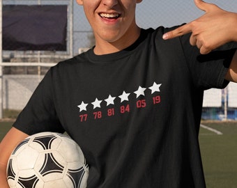 May include: A young person wearing a black t-shirt with white stars and the numbers 77 78 81 84 05 19 printed on it. They are holding a football and pointing to the shirt with their right hand.