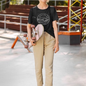 K&ouml;nnte beinhalten: Eine Person tr&auml;gt ein schwarzes T-Shirt mit einer wei&szlig;en Wellengrafik in einem Kreis. Das Shirt hat einen kleinen wei&szlig;en Mond und Sterne im Kreis. Die Person h&auml;lt ein Skateboard.