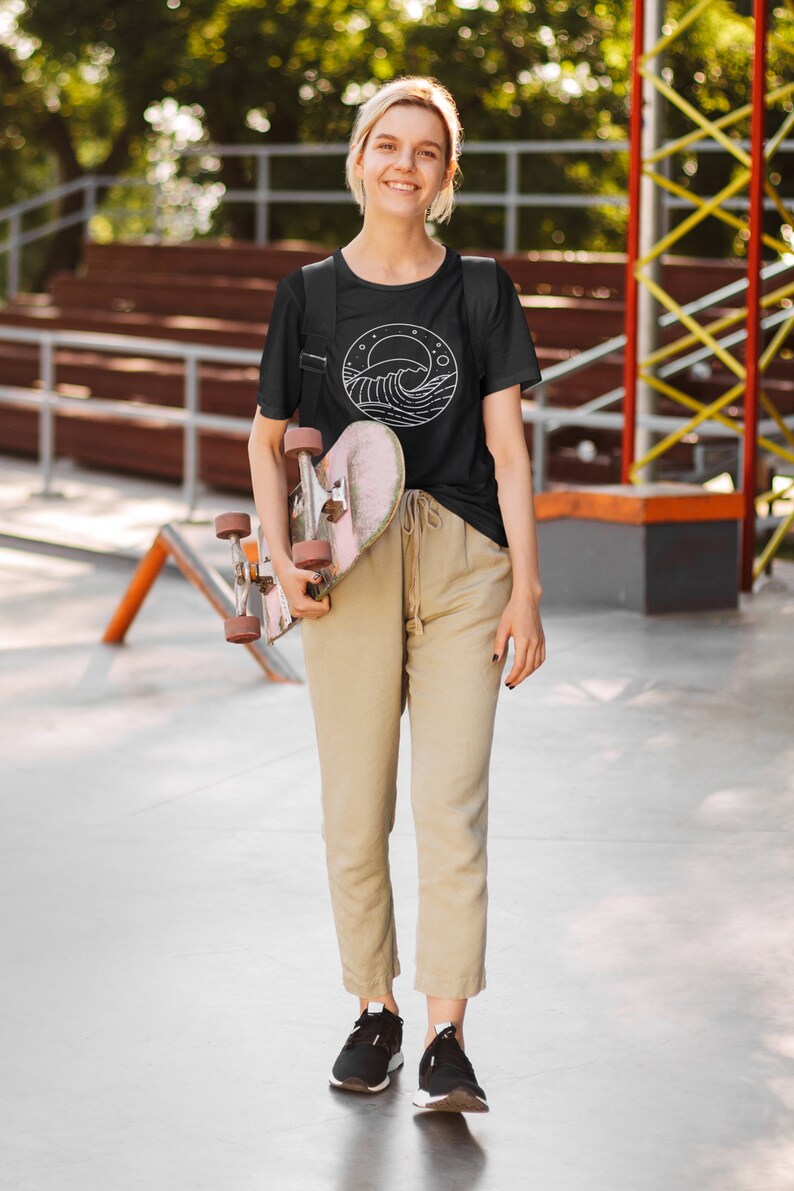 K&ouml;nnte beinhalten: Eine Person tr&auml;gt ein schwarzes T-Shirt mit einer wei&szlig;en Wellengrafik in einem Kreis. Das Shirt hat einen kleinen wei&szlig;en Mond und Sterne im Kreis. Die Person h&auml;lt ein Skateboard.