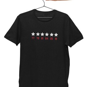 Pu&ograve; includere: Maglietta nera con cinque stelle bianche e i numeri 77 78 81 84 05 19 stampati in rosso sotto.