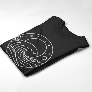 K&ouml;nnte beinhalten: Ein schwarzes T-Shirt mit einem wei&szlig;en Grafikdruck einer Welle in einem Kreis mit Sternen und Monden.