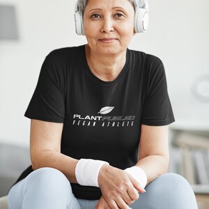 May include: A woman wearing a black t-shirt with the text "PLANT FUELED VEGAN ATHLETE" in white. She is sitting on a silver exercise ball and wearing white headphones.