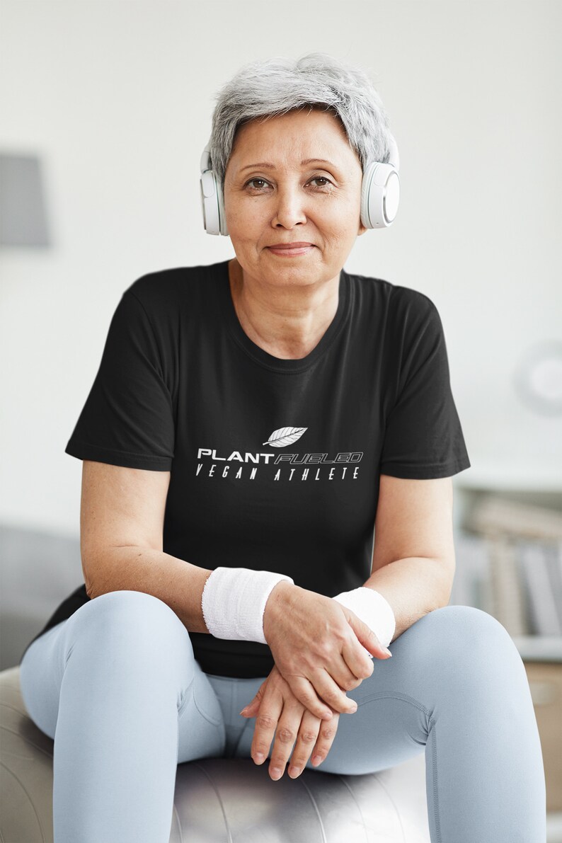 May include: A woman wearing a black t-shirt with the text "PLANT FUELED VEGAN ATHLETE" in white. She is sitting on a silver exercise ball and wearing white headphones.